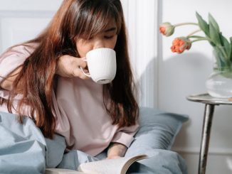 An Asian woman enjoying her morning with coffee and a book in a cozy bedroom setting.