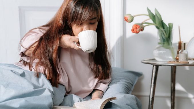 An Asian woman enjoying her morning with coffee and a book in a cozy bedroom setting.